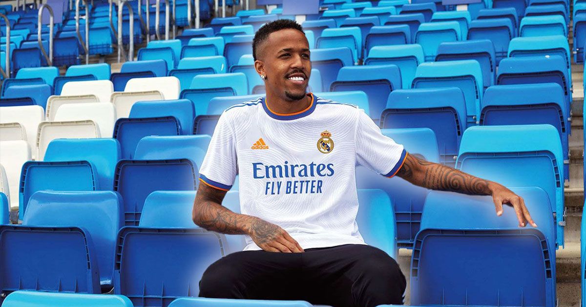 Éder Militão wearing a white Real Madrid kit featuring blue and orange branding while sat on a blue seat inside a stadium.