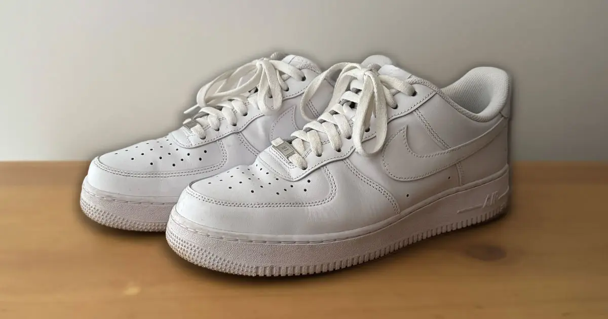 A pair of all-white Air Force 1 low-tops on a brown wooden surface.