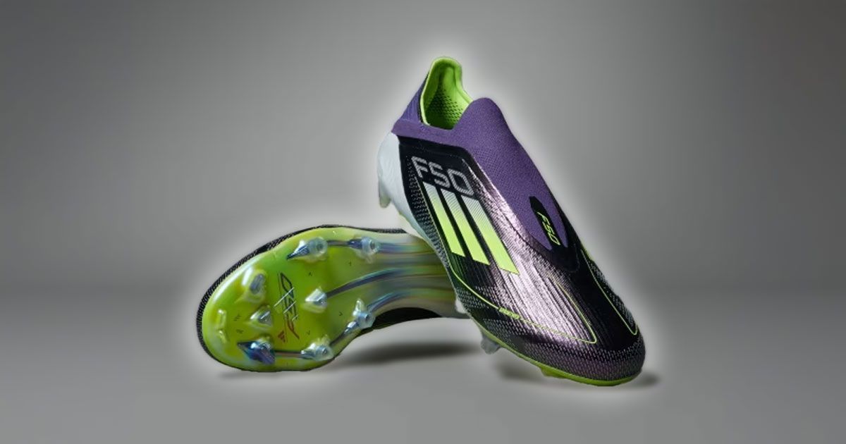 A purple, laceless football boot with lime green design details leaning against the other boot resting on its side.