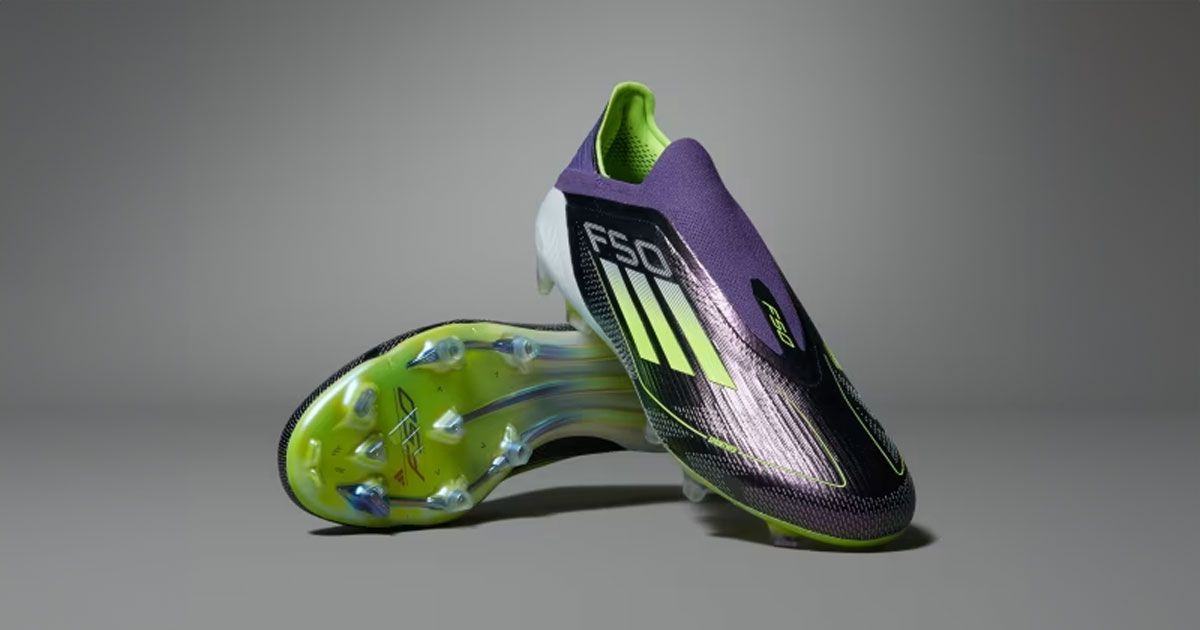 A purple, laceless football boot with lime green design details leaning against the other boot resting on its side.