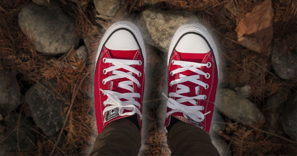 Someone standing on brown fallen leaves and rocks wearing a pair of red and white Converse Lows.