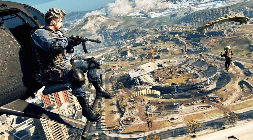 Warzone player holding a PPSH SMG while sitting on the outside of a helicopter flying above the Verdansk map. Players parachuting towards a stadium under construction are on the right-hand side