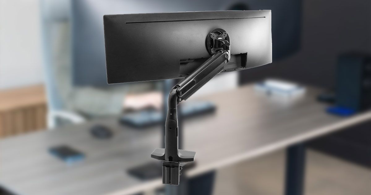 A black ultrawide monitor mounted to a monitor arm in front of a blurry image of someone sat at a mounted ultrawide monitor.
